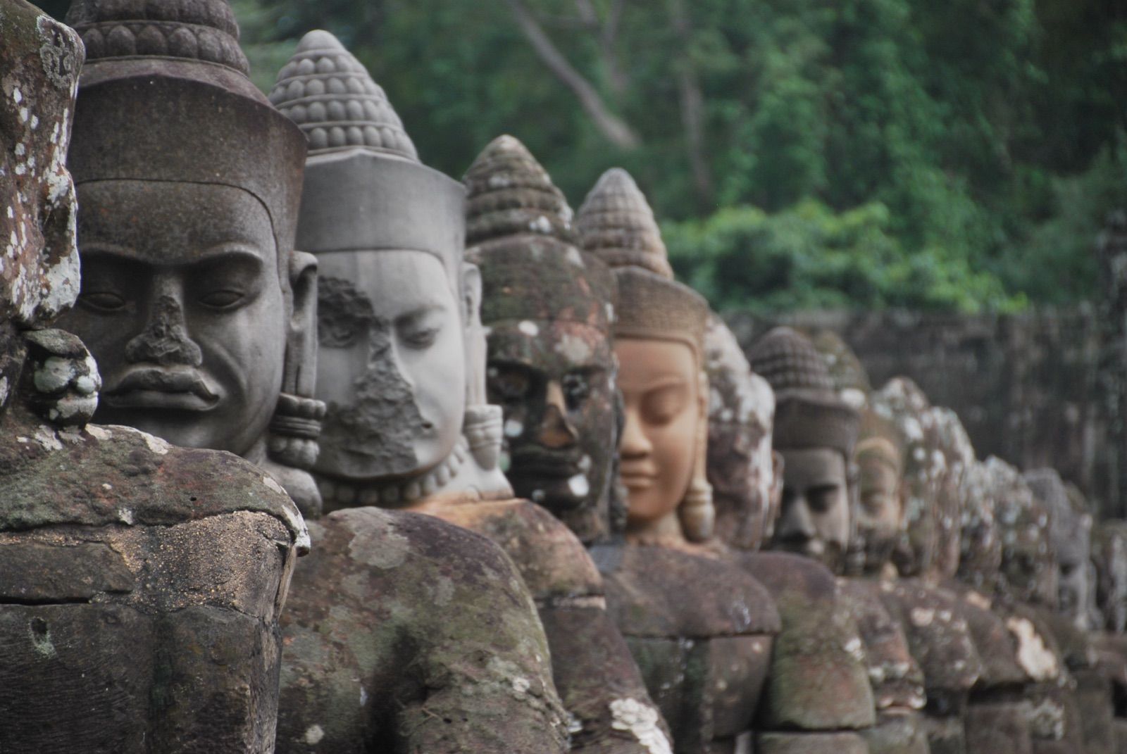 Hilera de cabezas de piedra de devas alineadas sobre la balaustrada de la puerta sur de Angkor Thom, con la selva desenfocada al fondo