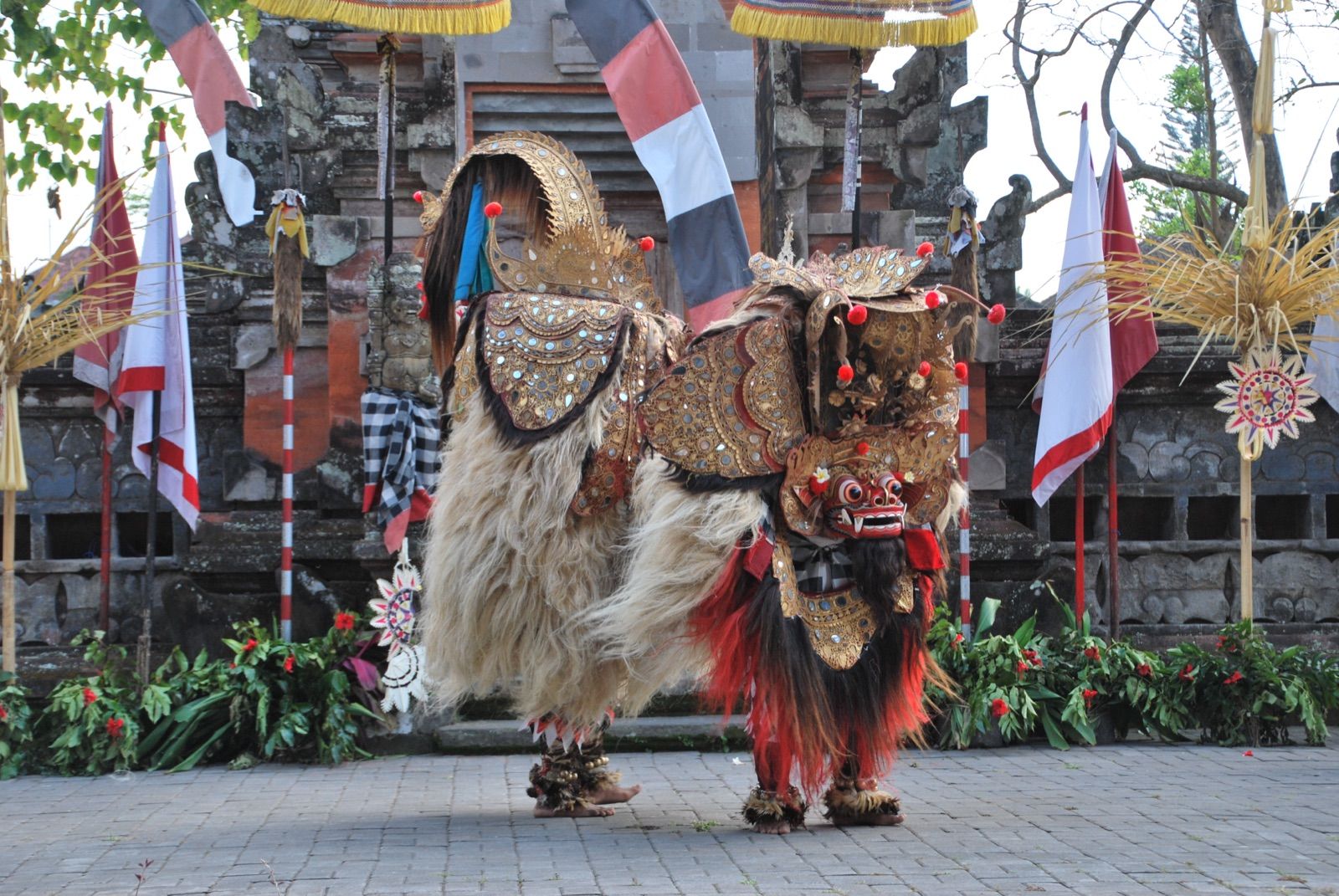 Figura completa del barong balinés, con melena blanca y armadura dorada de espejos, de pie en el patio empedrado de un templo entre banderas y ofrendas