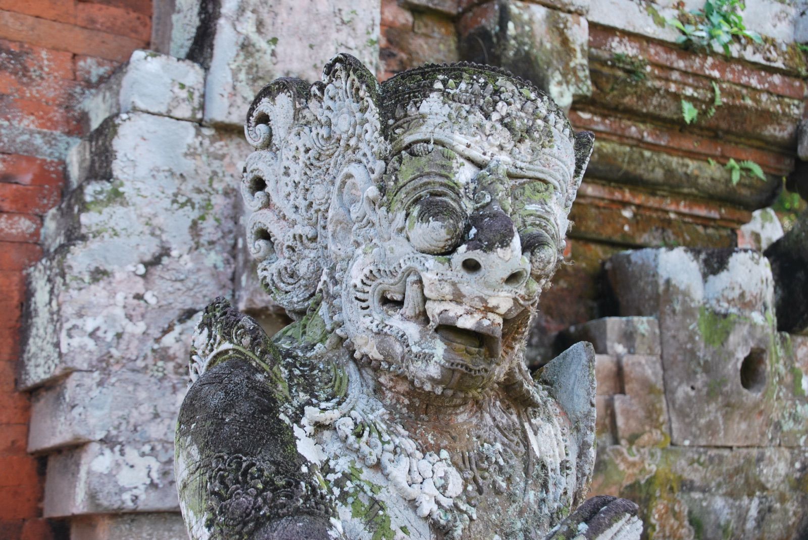 Estatua de guardián balinés tallada en piedra volcánica, cubierta de líquenes, ante un muro de ladrillo rojo de un templo