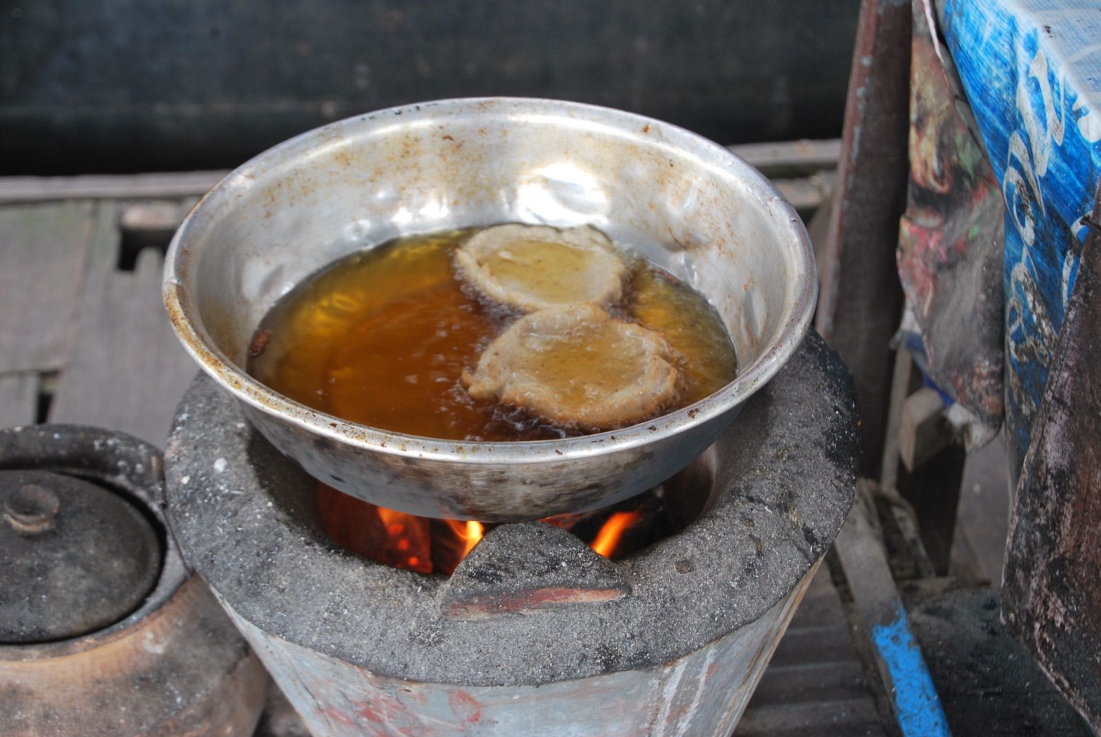 Cuenco metálico abollado con dos buñuelos friéndose en aceite sobre un brasero de carbón encendido en un puesto de comida de carretera