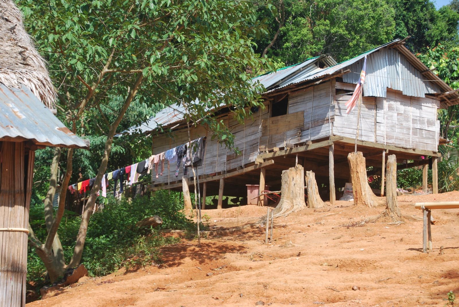 Casa de madera sobre pilotes con tejado de chapa y ropa tendida en un cordel, en un claro de tierra roja rodeado de selva