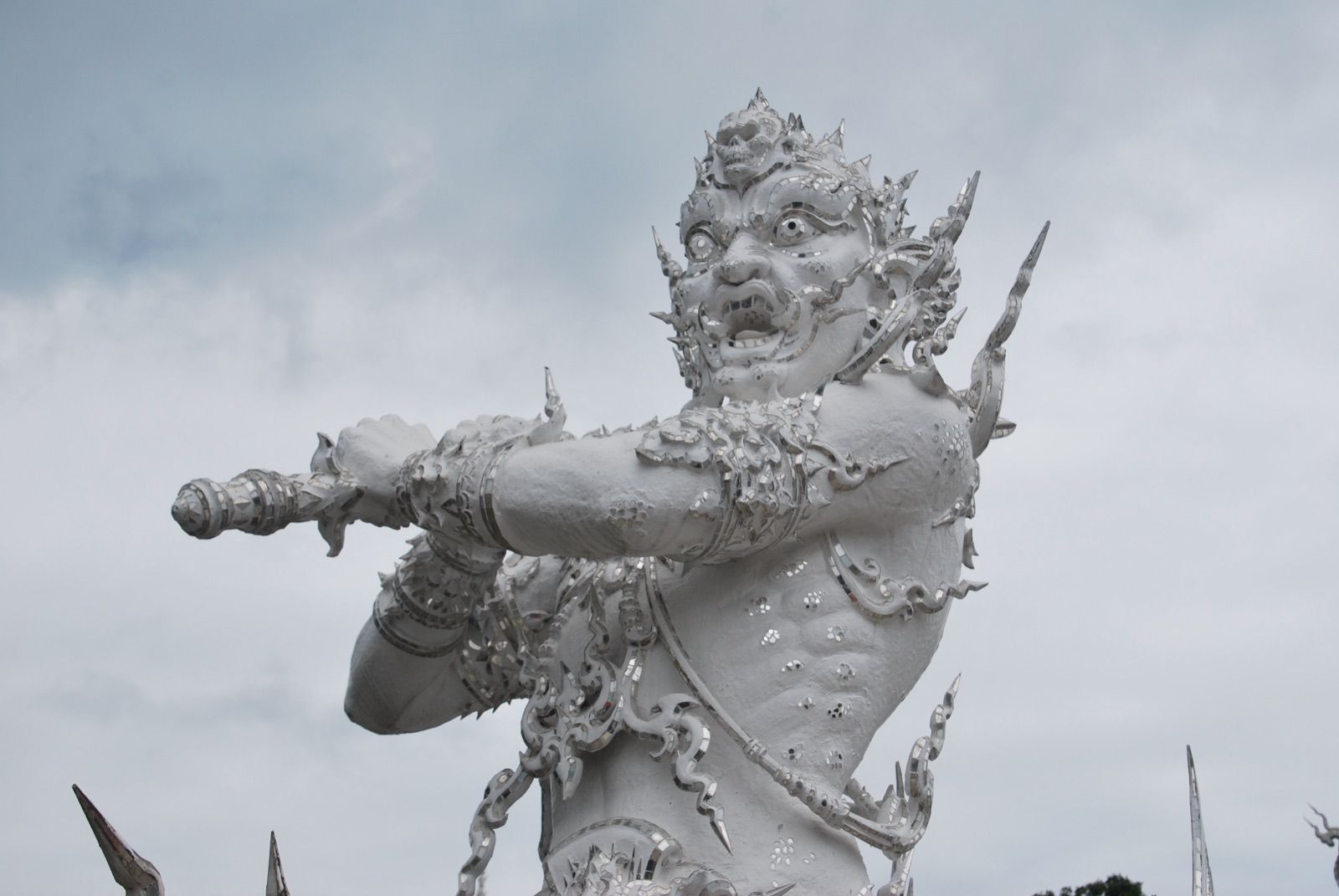 Estatua blanca de un demonio con colmillos y ojos saltones blandiendo una maza hacia el frente, con el cielo nublado de fondo