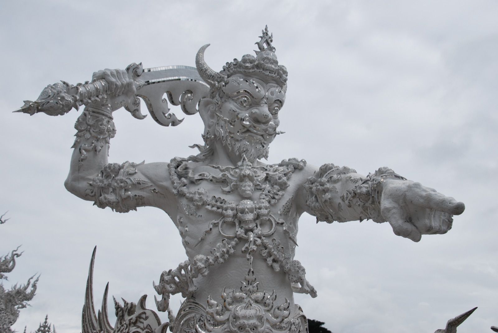 Escultura blanca de un guardián demoníaco con cuernos, brazos extendidos y collar de calaveras, recubierta de mosaico de espejo, recortada contra un cielo encapotado
