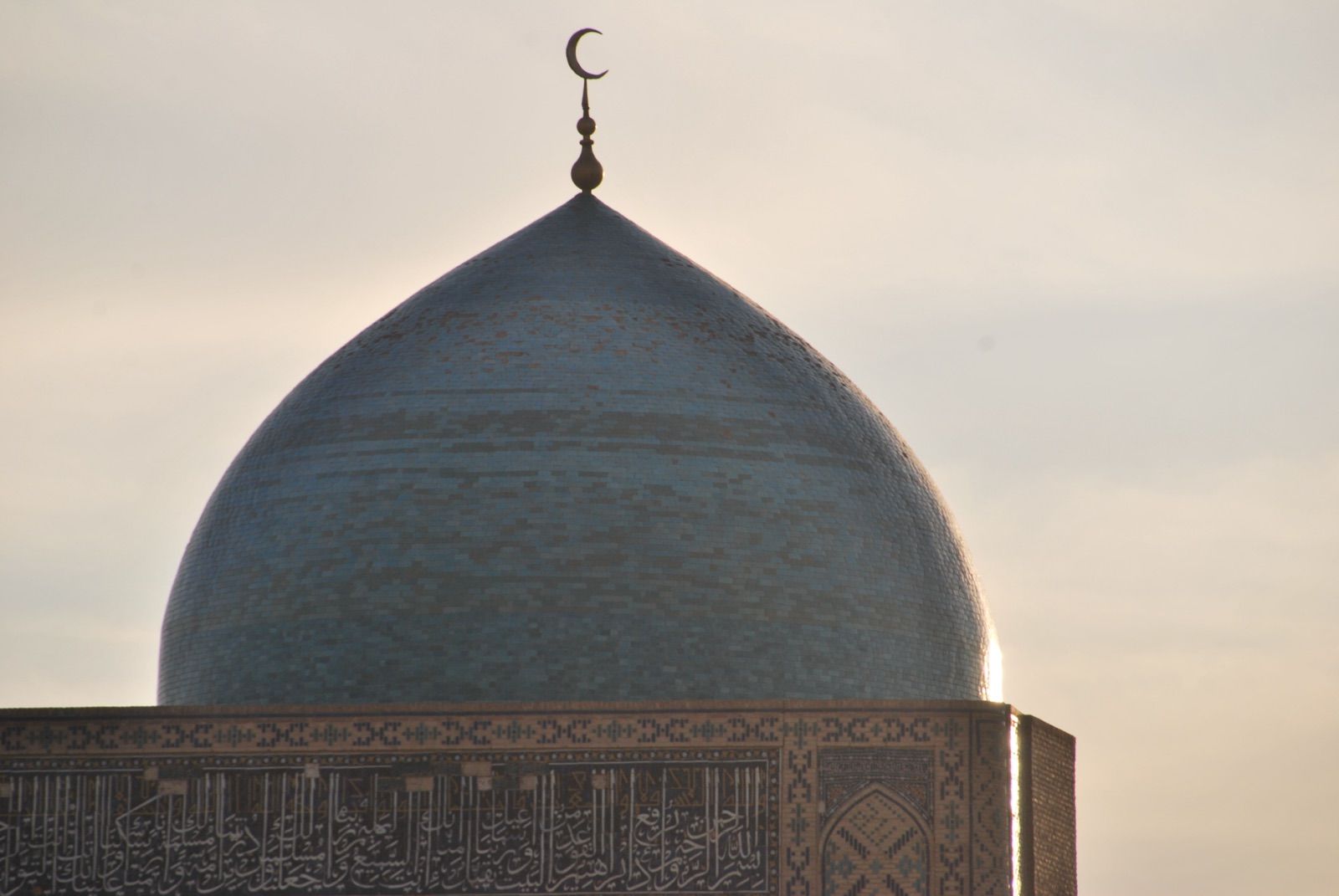 Cúpula turquesa rematada por una media luna metálica sobre un tambor cubierto de caligrafía en azulejo, recortada a contraluz contra un cielo pálido