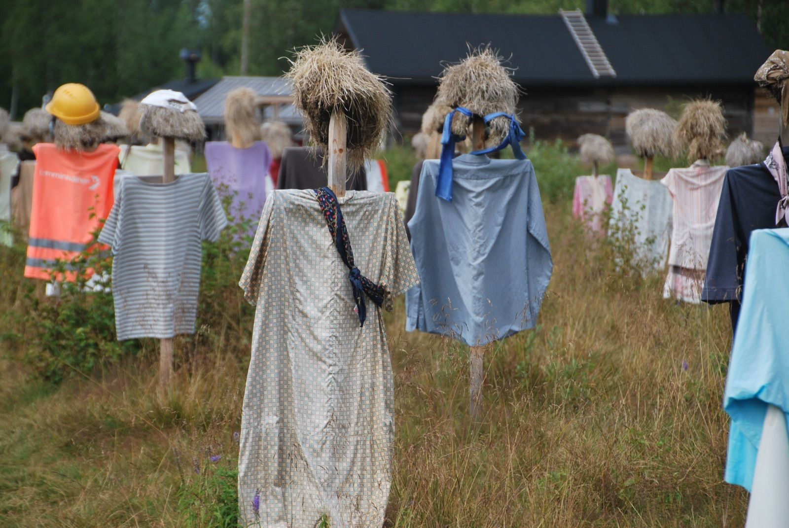 Dos figuras vestidas con bata y chubasquero azul, con cabezas de hierba seca, ante un granero de madera oscura con tejado negro en un campo de hierba alta