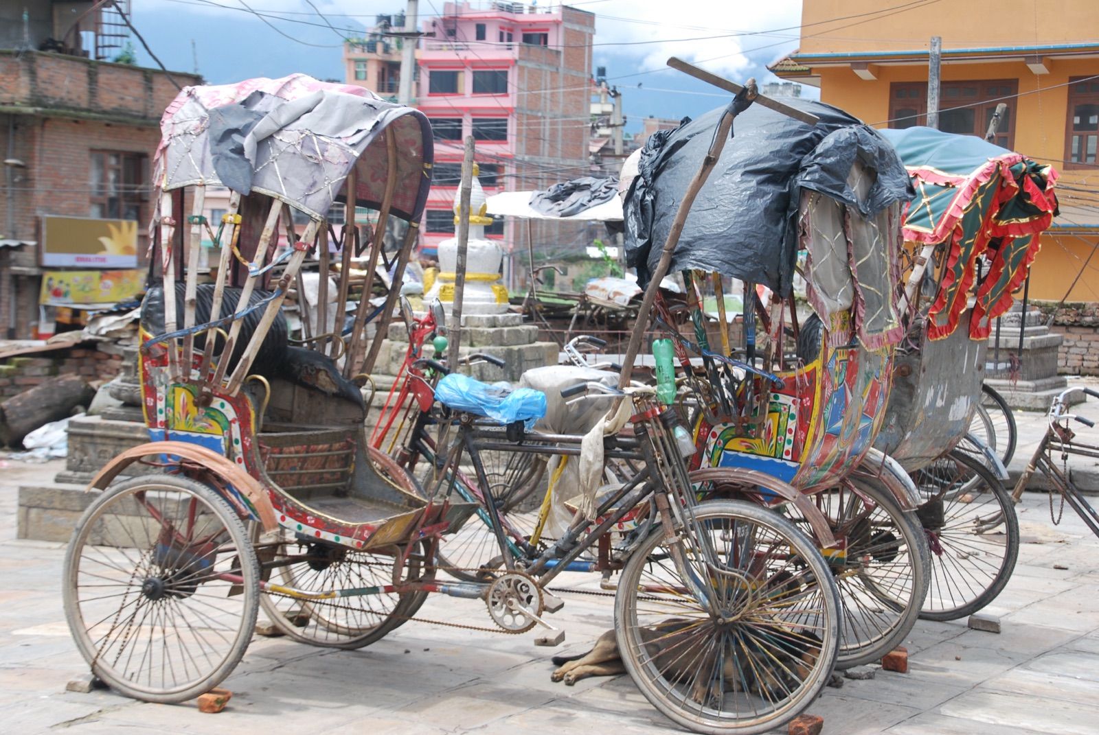 Varios rickshaws de pedales con toldos remendados y ruedas oxidadas aparcados en una plaza de tierra