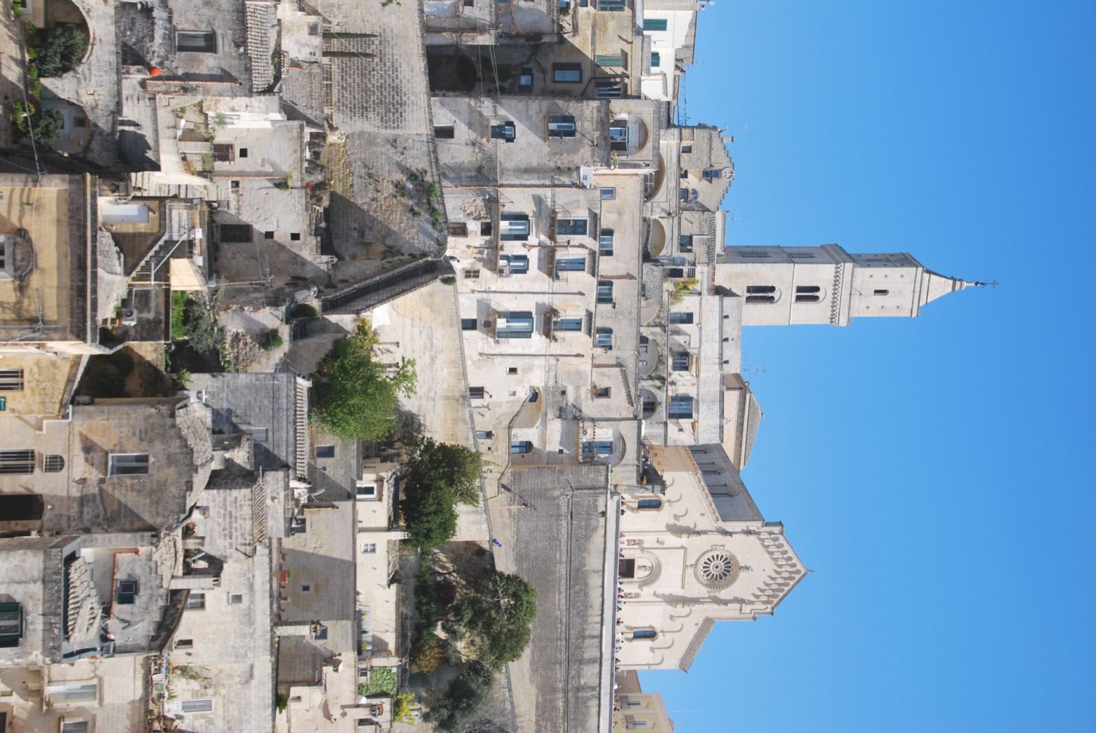 Vista vertical de los Sassi de Matera, con casas de piedra escalonadas y el campanario de la catedral recortado contra un cielo azul limpio