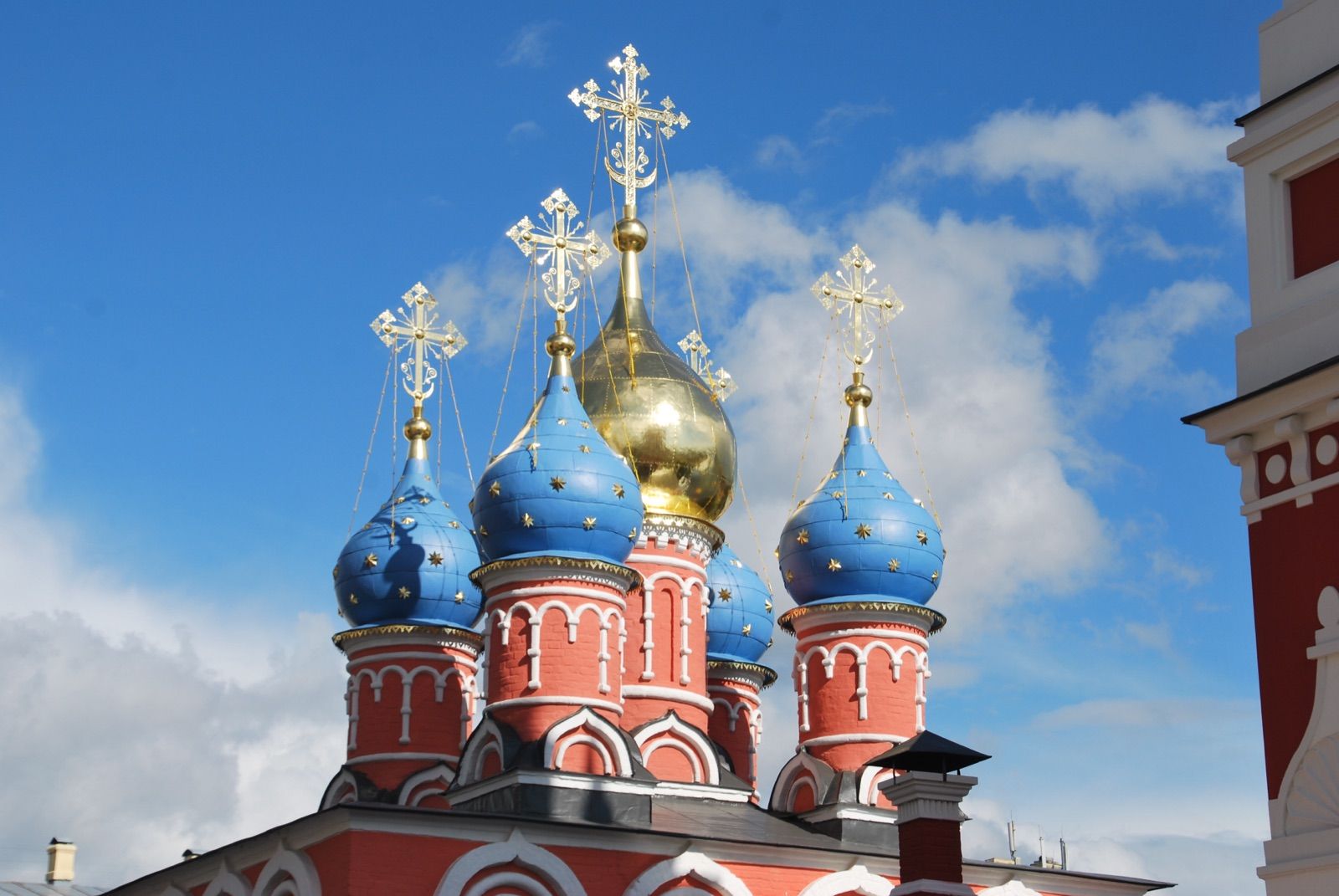 Cinco cúpulas bulbosas de una iglesia ortodoxa, cuatro azules con estrellas doradas y una central dorada, con cruces y cadenas recortadas contra un cielo azul con nubes blancas