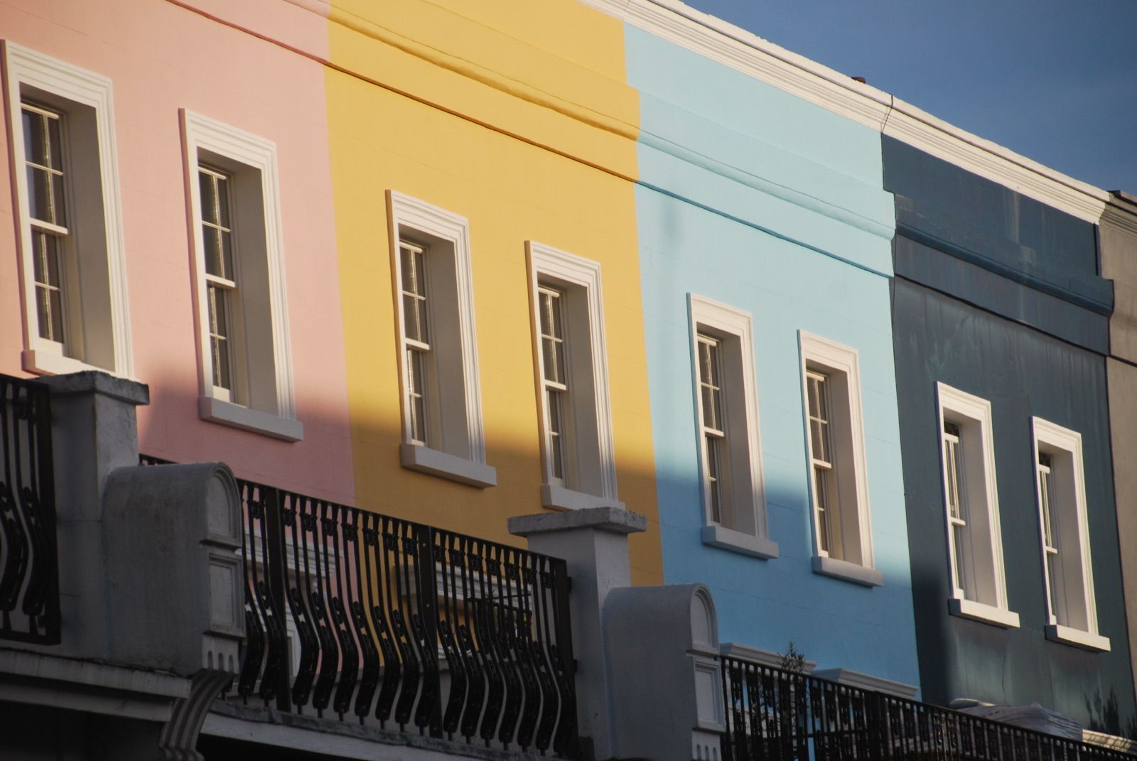 Hilera de casas adosadas de Notting Hill con fachadas pintadas en rosa, amarillo, azul claro y gris pizarra, ventanas de marco blanco y una sombra diagonal cruzando el conjunto