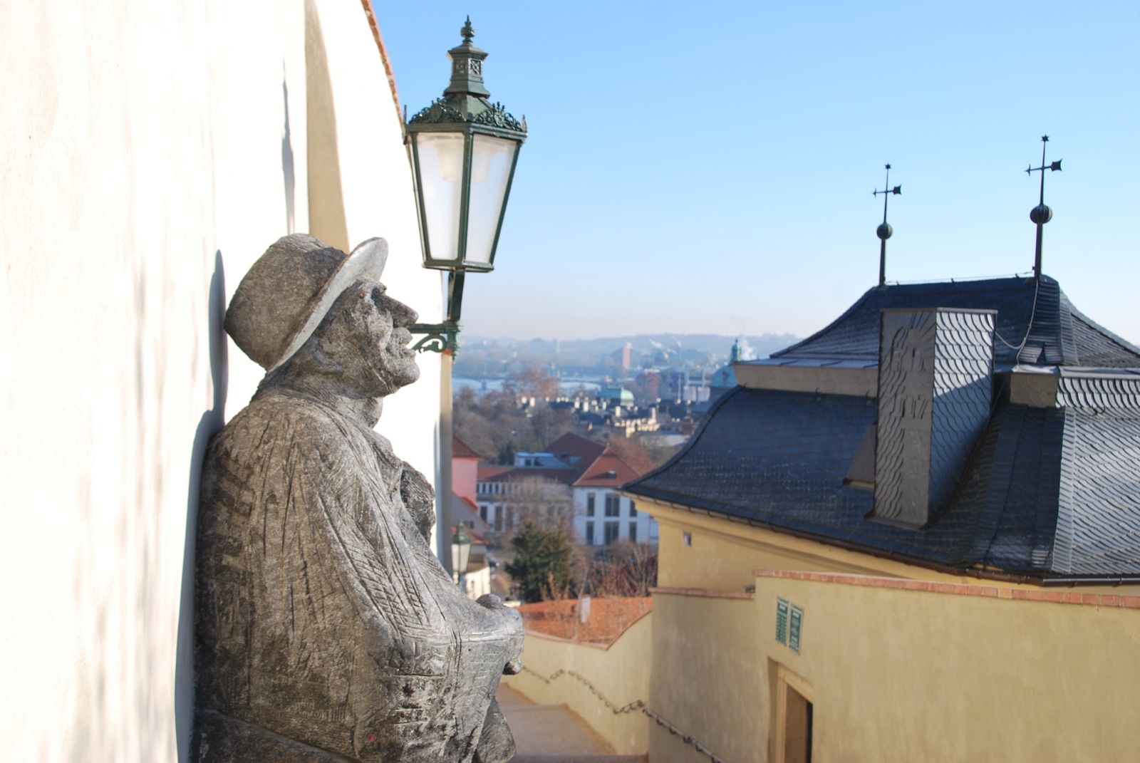 Estatua de piedra de un hombre sentado con sombrero junto a una farola clásica en un mirador de Praga, con tejados de pizarra y el río Moldava con sus puentes entre la bruma al fondo