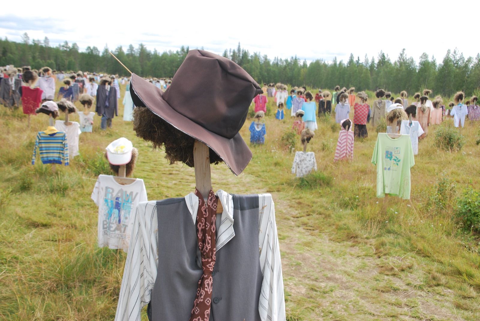 Figura de espantapájaros con sombrero marrón, chaleco y corbata en primer plano, y un sendero de tierra que atraviesa un campo con cientos de figuras vestidas hasta el bosque
