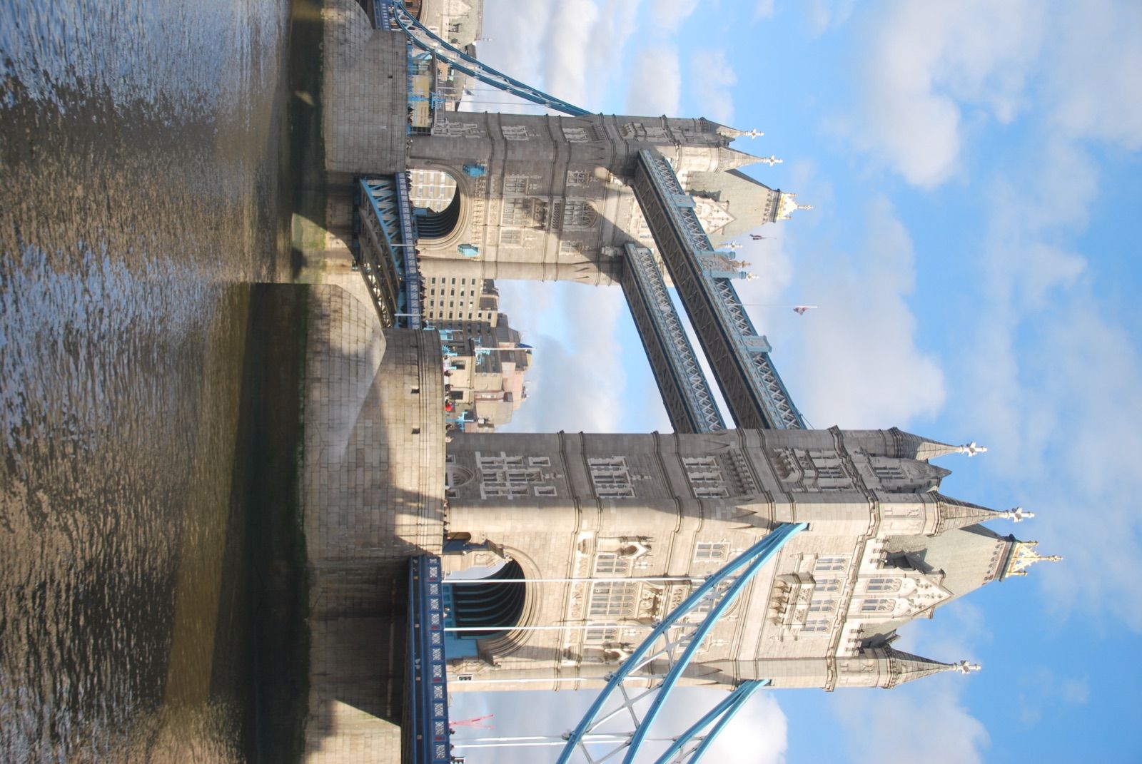 Tower Bridge en vertical visto desde la orilla del Támesis, con las dos torres de piedra, los tirantes azules y cielo azul con nubes blancas