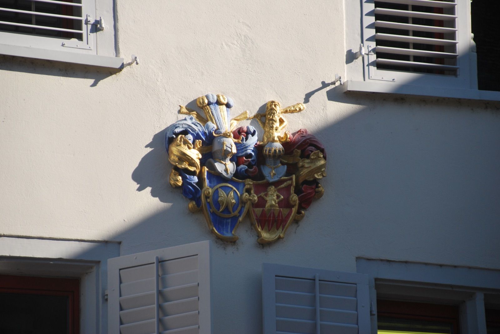 Escudo de armas doble policromado en azul, rojo y pan de oro montado sobre una fachada blanca, entre ventanas con contraventanas de lamas