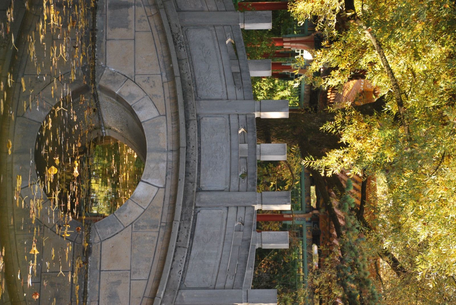 Puente peatonal de piedra con balaustrada tallada sobre un estanque cubierto de hojas amarillas, con farolillos rojos y un pabellón chino entre los árboles en otoño