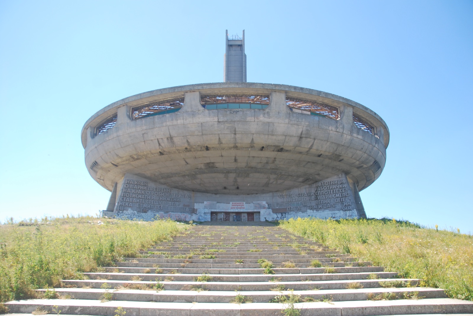 Monumento de Buzludzha visto de frente: el platillo de hormigón y su torre sobre una escalinata invadida por la hierba