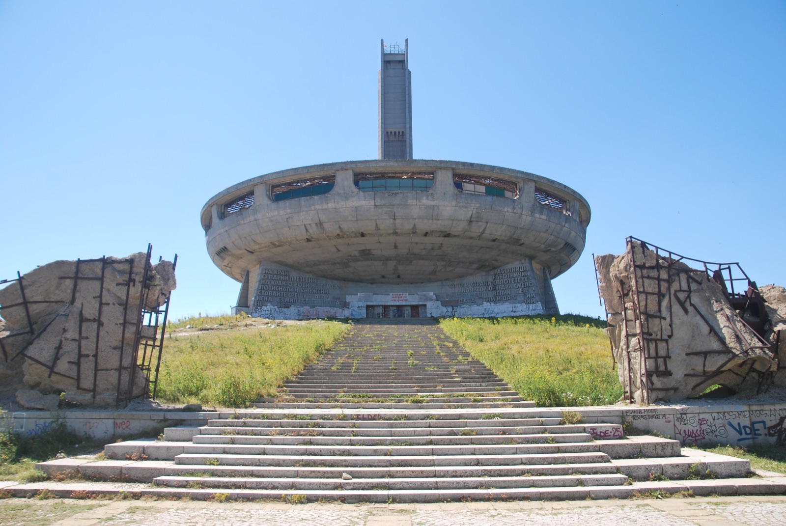 Monumento de Buzludzha enmarcado entre las esculturas rotas de la entrada, con las escaleras cubiertas de vegetación