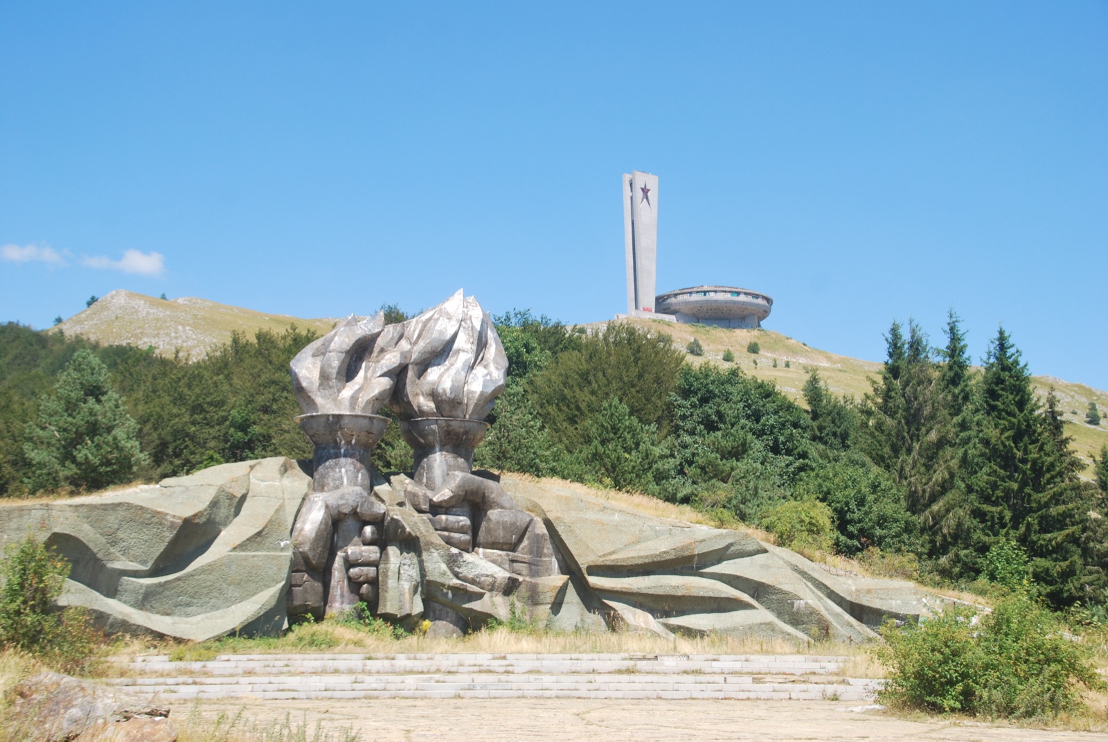 Escultura de dos antorchas de hormigón al pie del monte, con el monumento de Buzludzha y su torre con estrella roja al fondo