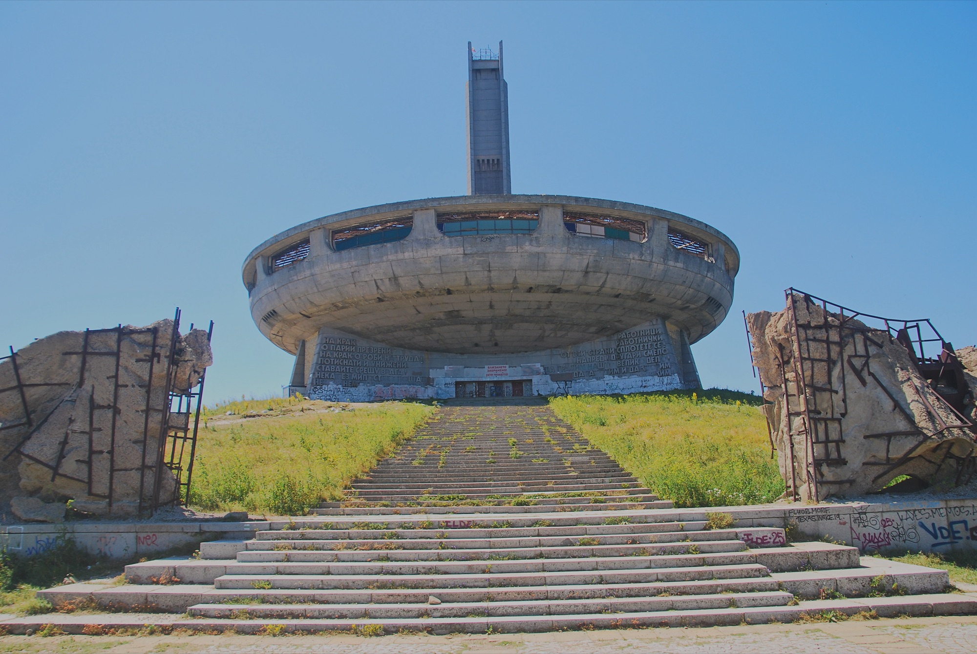 Monumento de Buzludzha con su platillo de hormigón y la torre de la estrella roja, visto de frente entre las puertas escultóricas en ruinas y la escalinata invadida de hierba