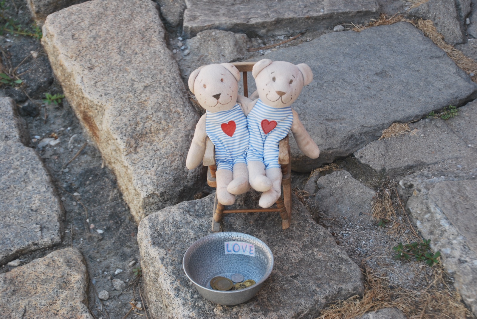 Dos ositos de trapo sentados en una silla en miniatura sobre piedras antiguas, con un cuenco de monedas rotulado LOVE