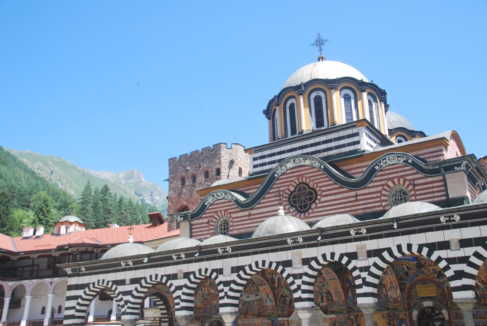 Iglesia principal del monasterio de Rila con su cúpula y arcos a rayas blancas y negras, la torre de Hrelyo y las montañas al fondo
