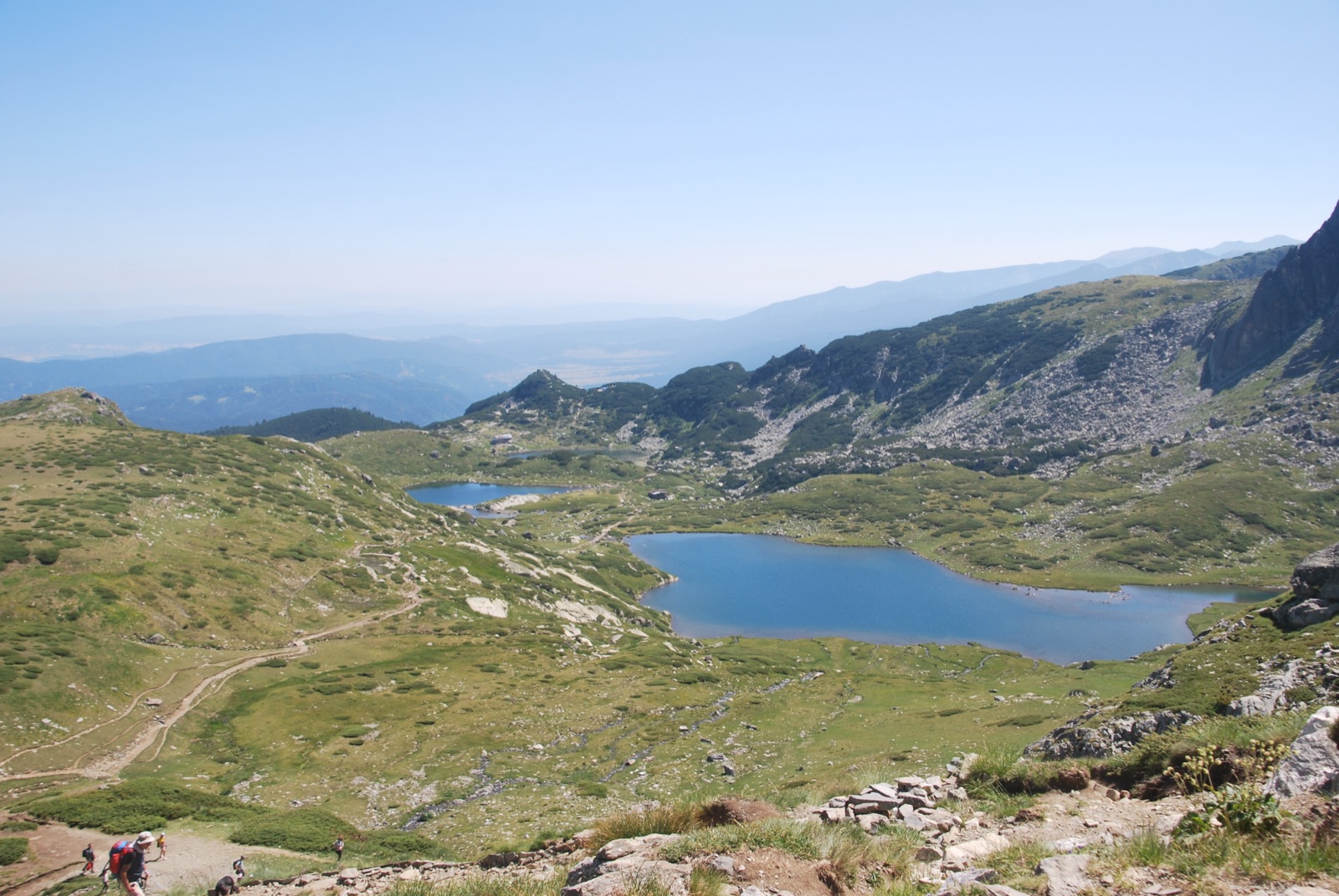 Panorámica de los Siete Lagos de Rila desde lo alto del sendero, con dos lagos azules en el valle y senderistas de espaldas en primer término