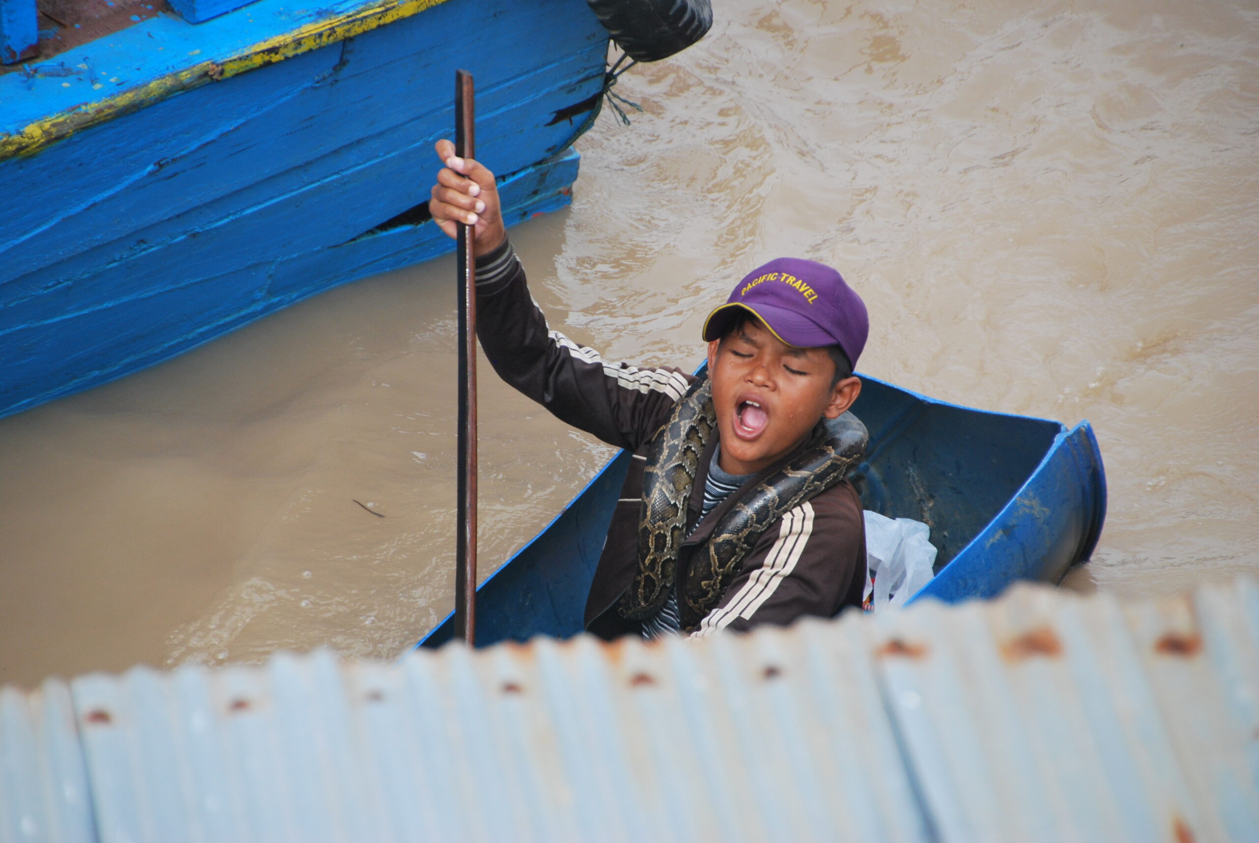 Niño camboyano con pitón enrollada al cuello remando junto a una barca azul en Tonlé Sap