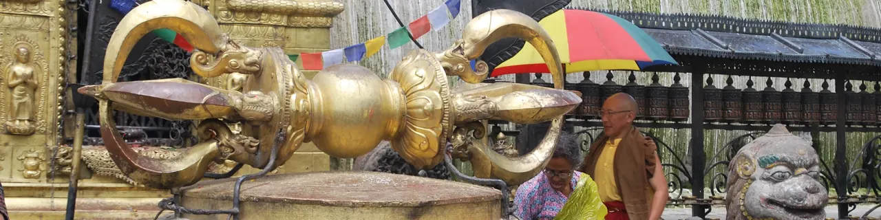 Cetro vajra dorado sobre un pedestal, con banderas de oración y un león de piedra al fondo